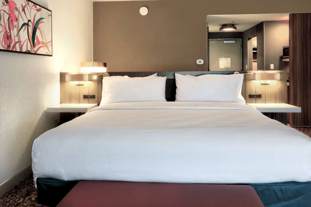 Hotellihuone, jossa on suuri sänky, kaksi yöpöytää, yölamppu, peili ja seinään kiinnitetty valaisin.