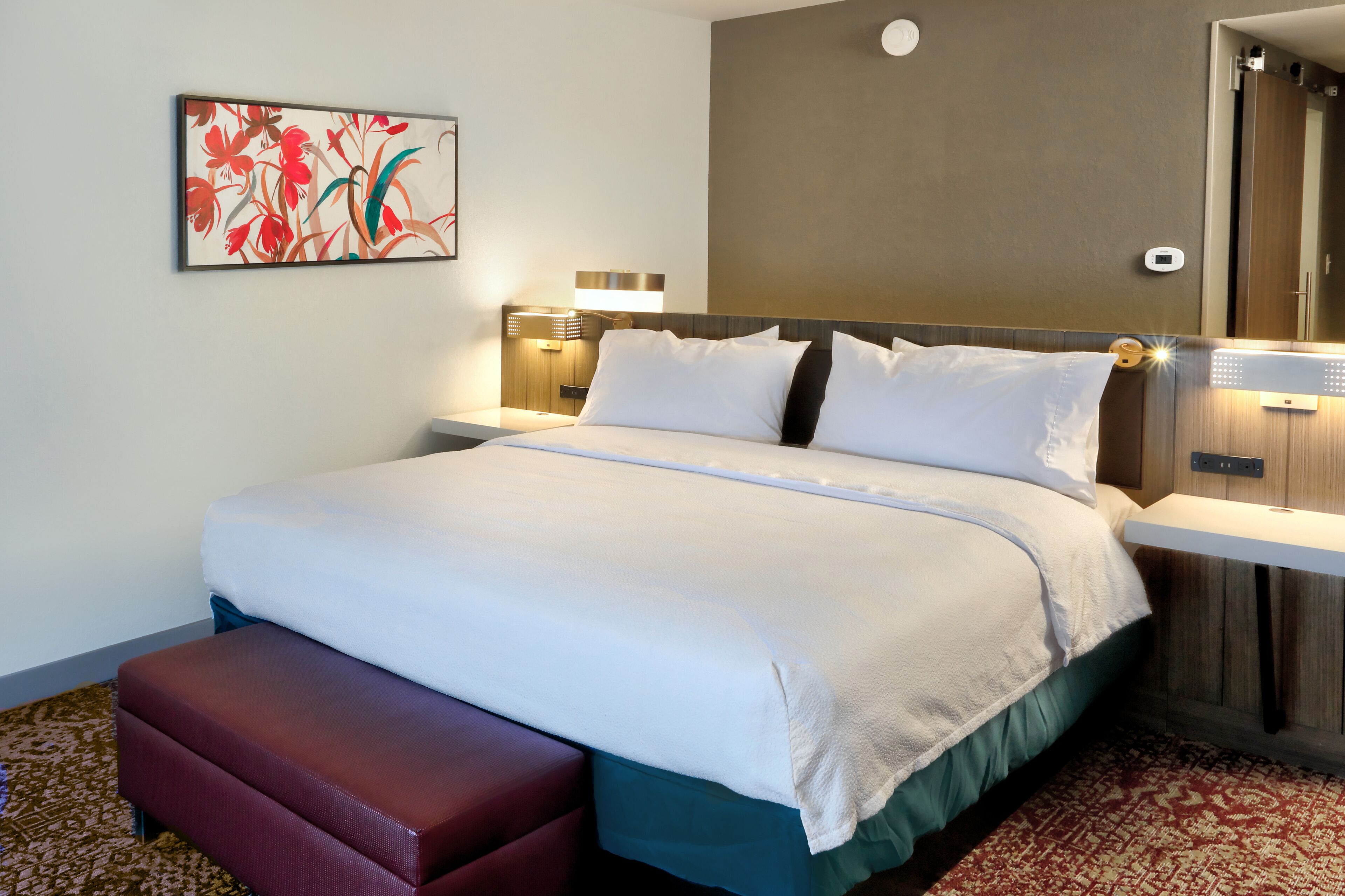 A hotel room with a large bed, a bench, a nightstand with a lamp, and a wall-mounted artwork.
