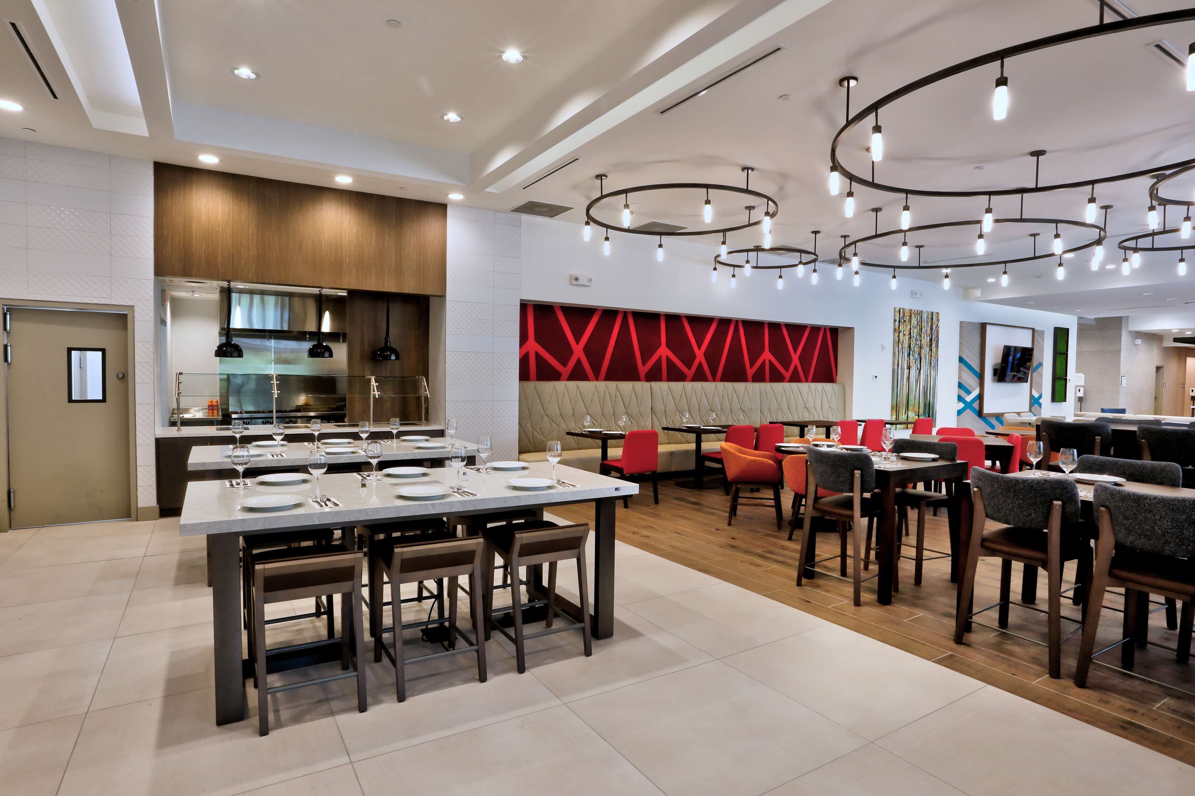 A modern restaurant with a central dining area, a kitchen, and a seating area with red chairs.