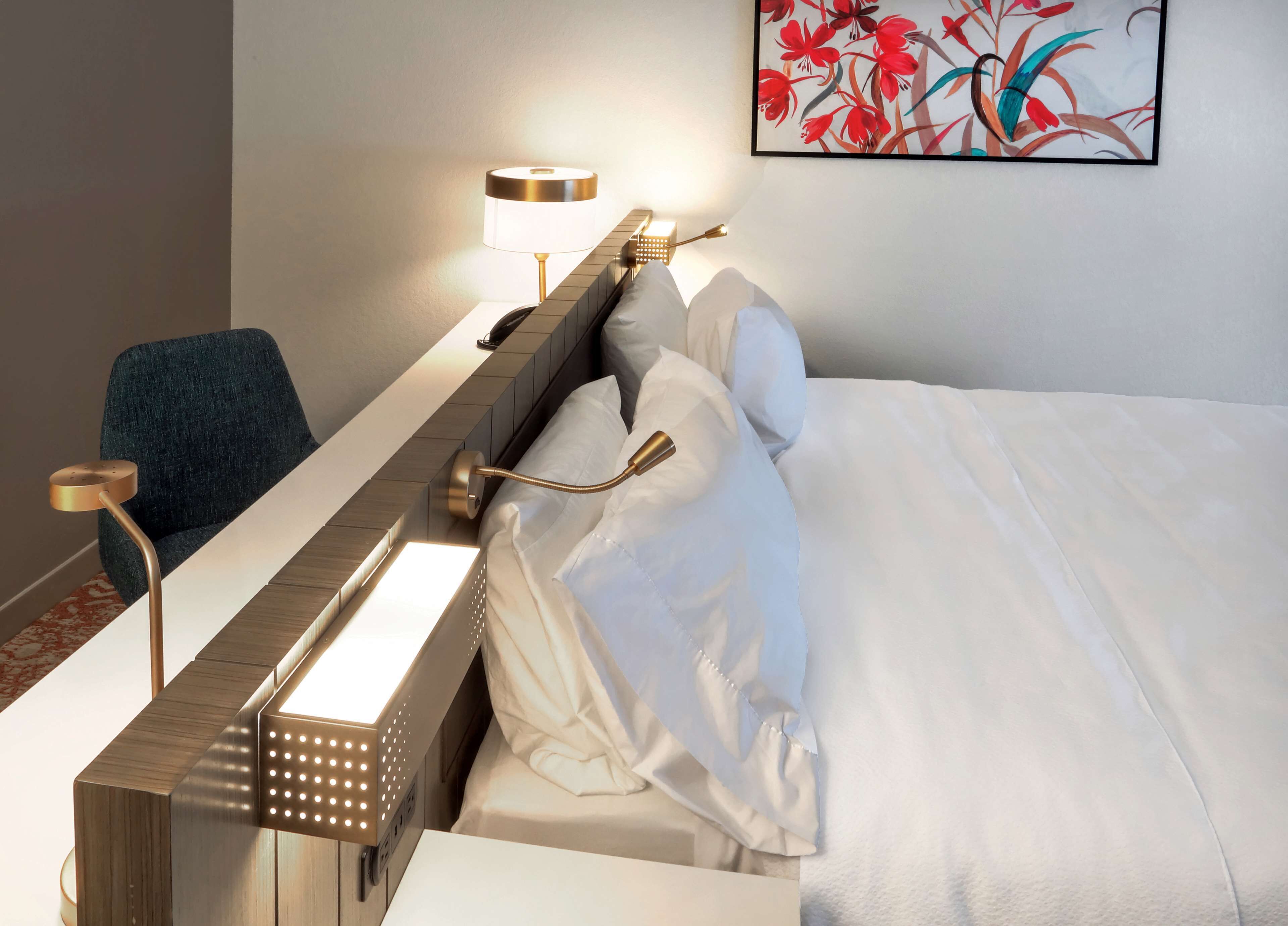 A hotel room with a neatly made bed, a bedside table with a lamp, a chair, and a wall-mounted phone.