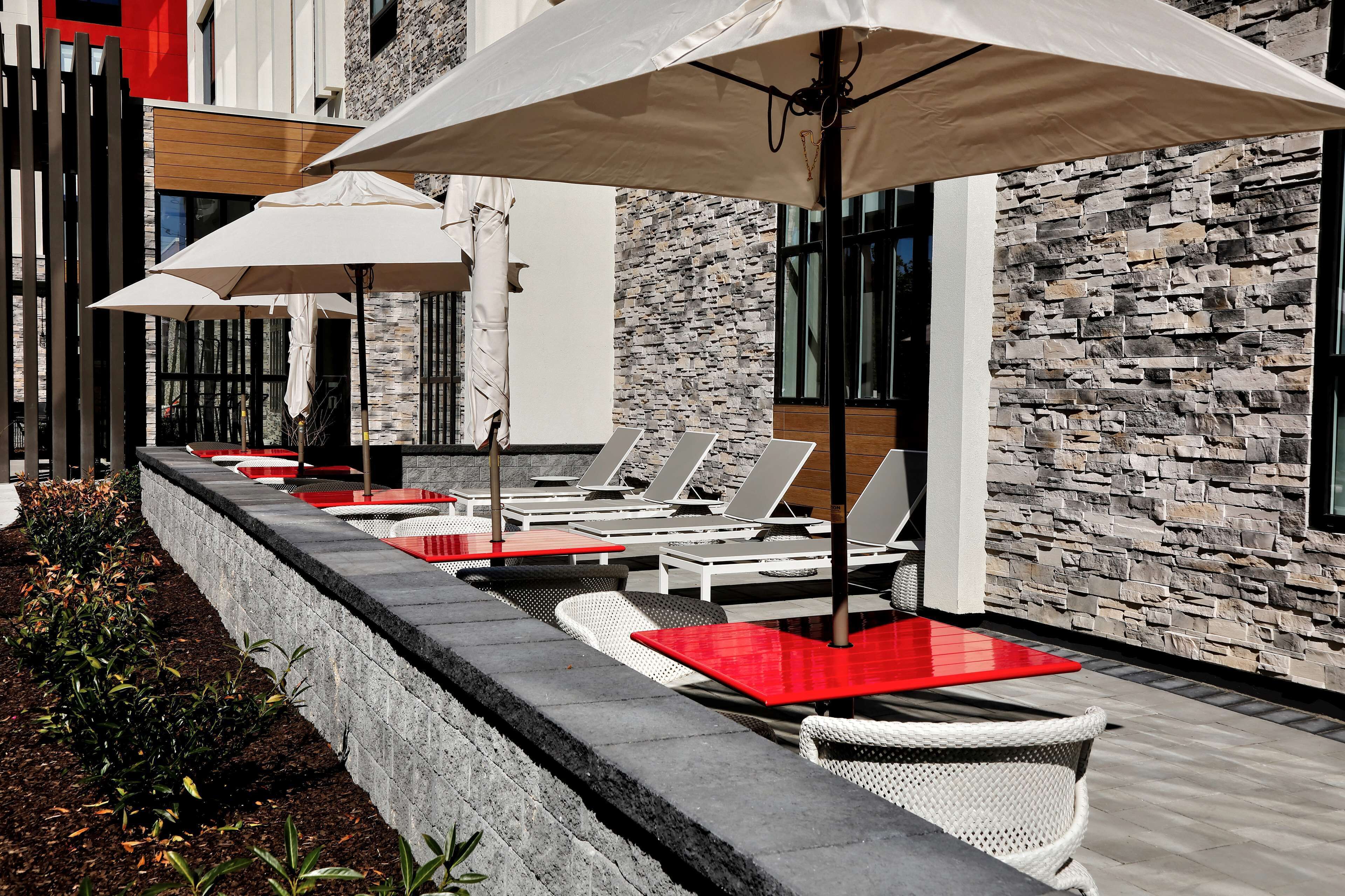 A row of sun loungers with red cushions and white umbrellas, set against a stone wall with a modern building in the background.