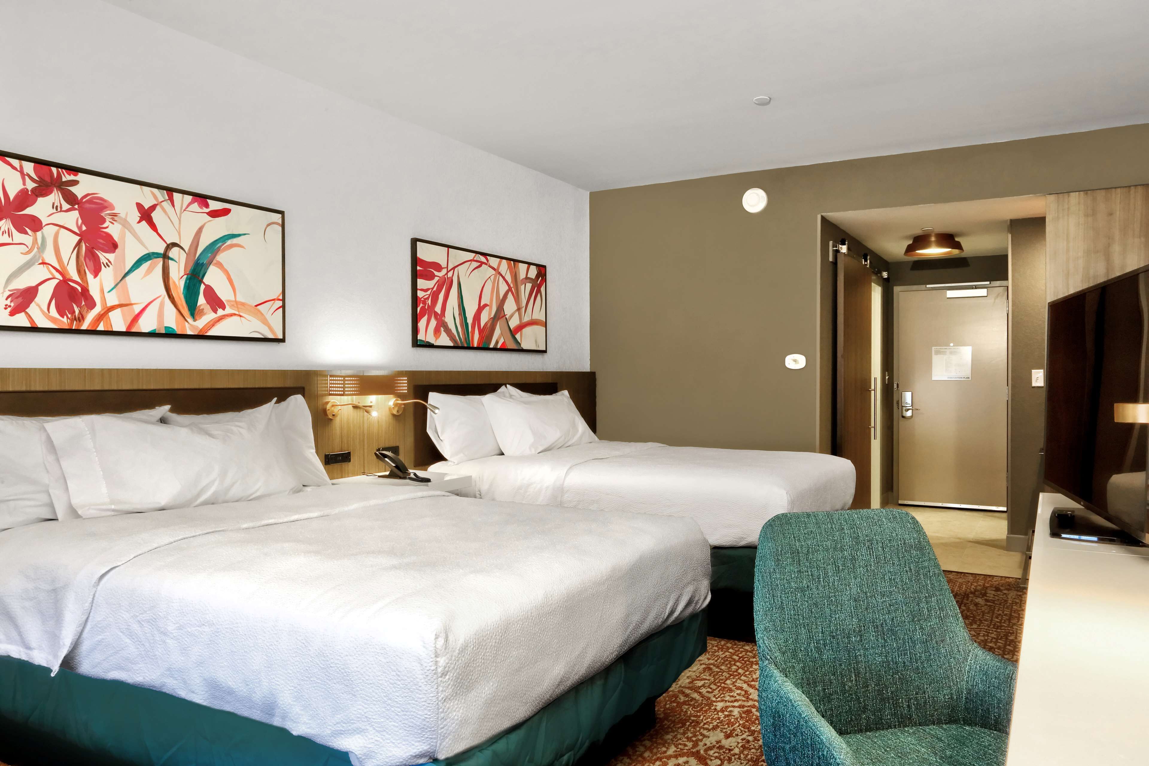 Habitación de hotel con dos camas, un sillón verde y dos cuadros con motivos florales.