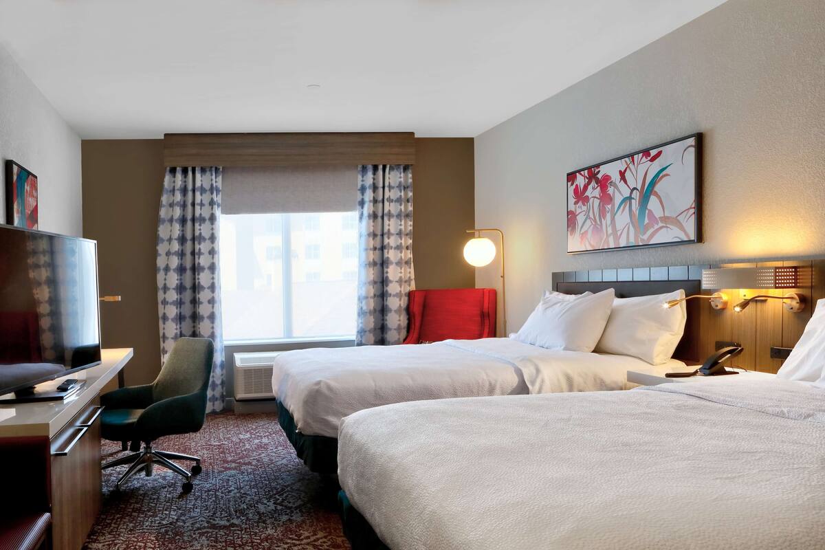 Hotellihuone, jossa on kaksi sänkyä, työpöytä tuolilla, televisio ja suuri ikkuna, jossa on verhot.