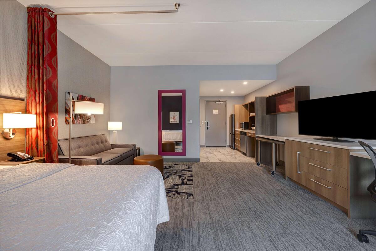 A hotel room with a bed, bedside lamps, a sofa, a desk, a television, and a kitchenette.