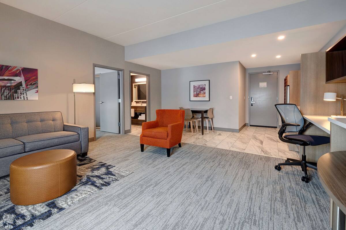 A modern hotel room with a grey sofa, an orange armchair, a round ottoman, a desk with a chair, and a large painting on the wall.