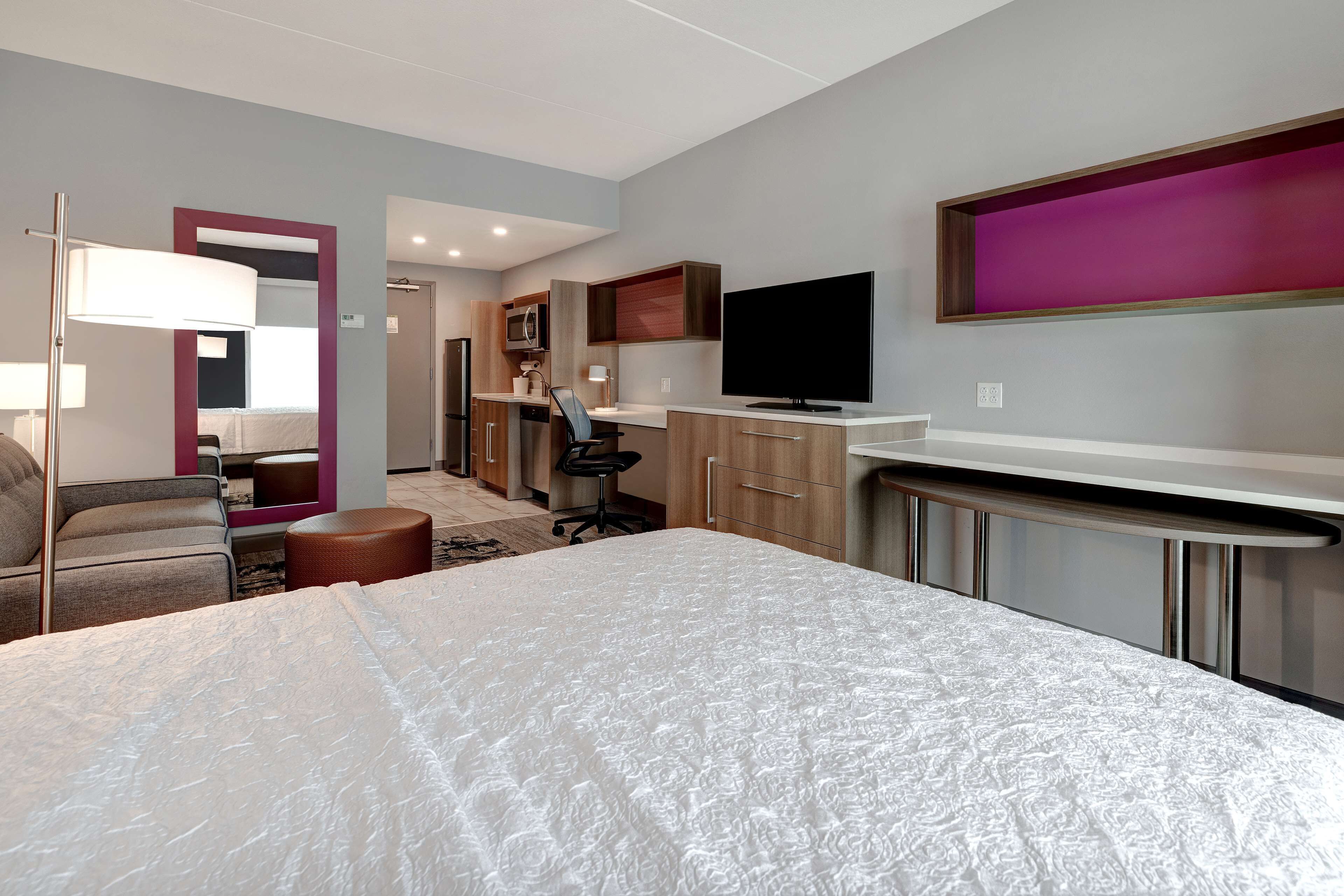 Quarto de hotel com uma cama grande, uma mesa de trabalho, televisão de tela plana, cozinha compacta e banheiro.