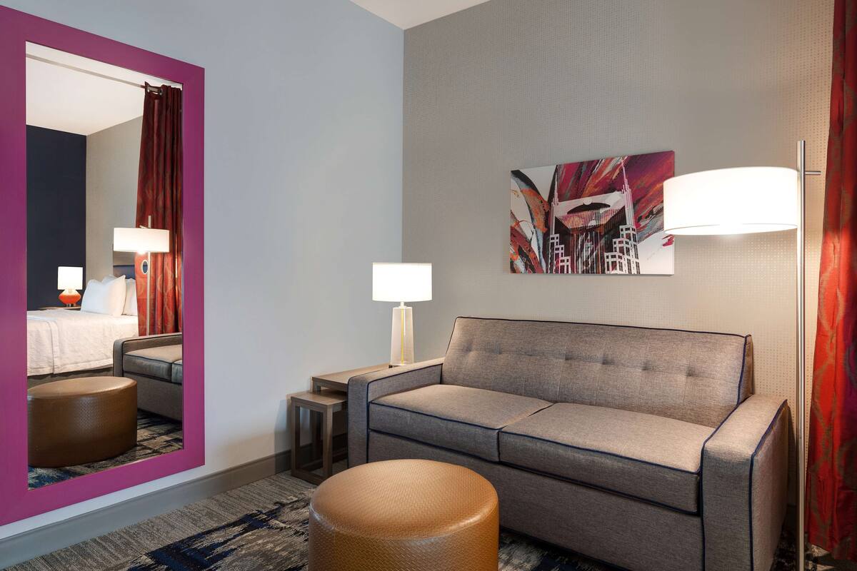 A hotel room with a grey sofa, a round ottoman, a small side table, a wall-mounted lamp, and a painting.