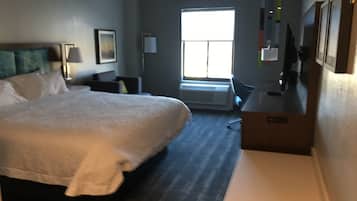 A hotel room with a large bed, a desk, a chair, a TV, and a window with blinds.