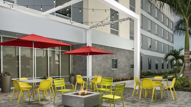 An outdoor patio with yellow chairs and red umbrellas, a fire pit, and a modern building in the background.