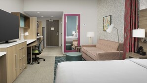 A hotel room with a bed, a desk with a computer, a chair, a sofa, a mirror, and a nightstand with a lamp.