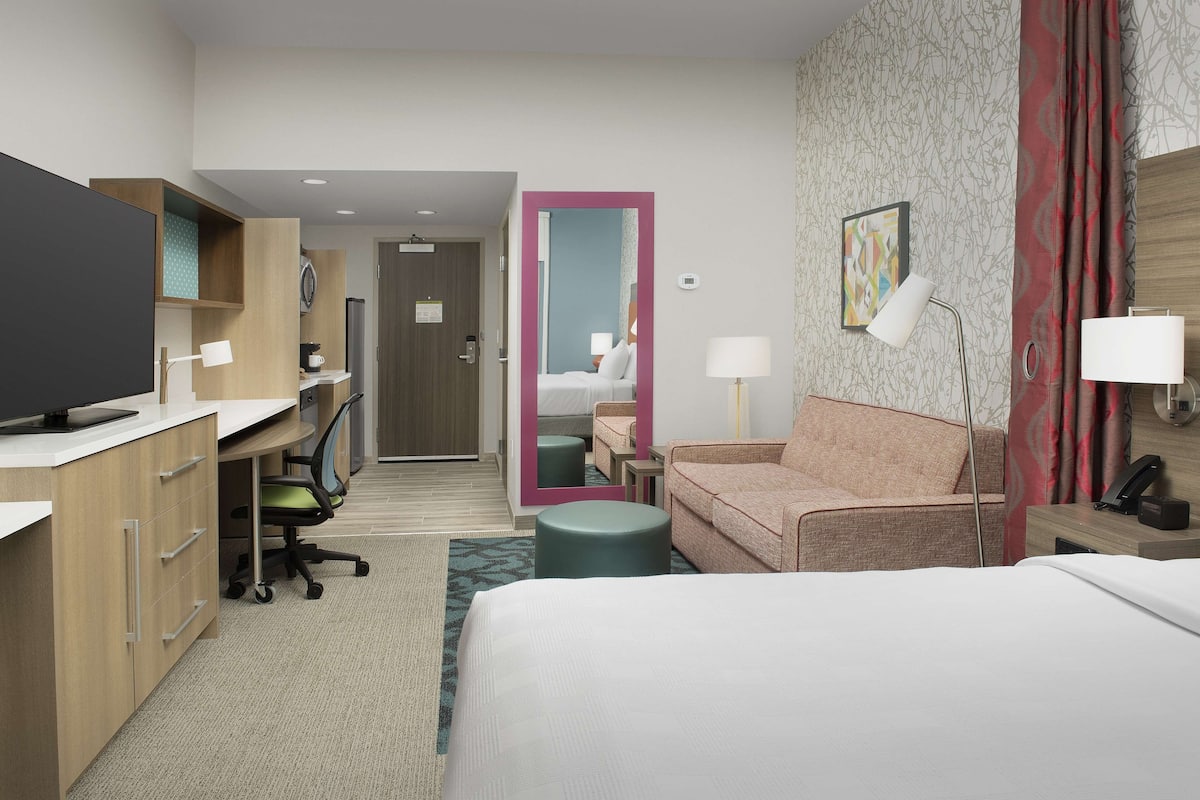 Hotelkamer met een bed, een bureau met computer, een stoel, een bank, een spiegel en een nachtkastje met lamp.