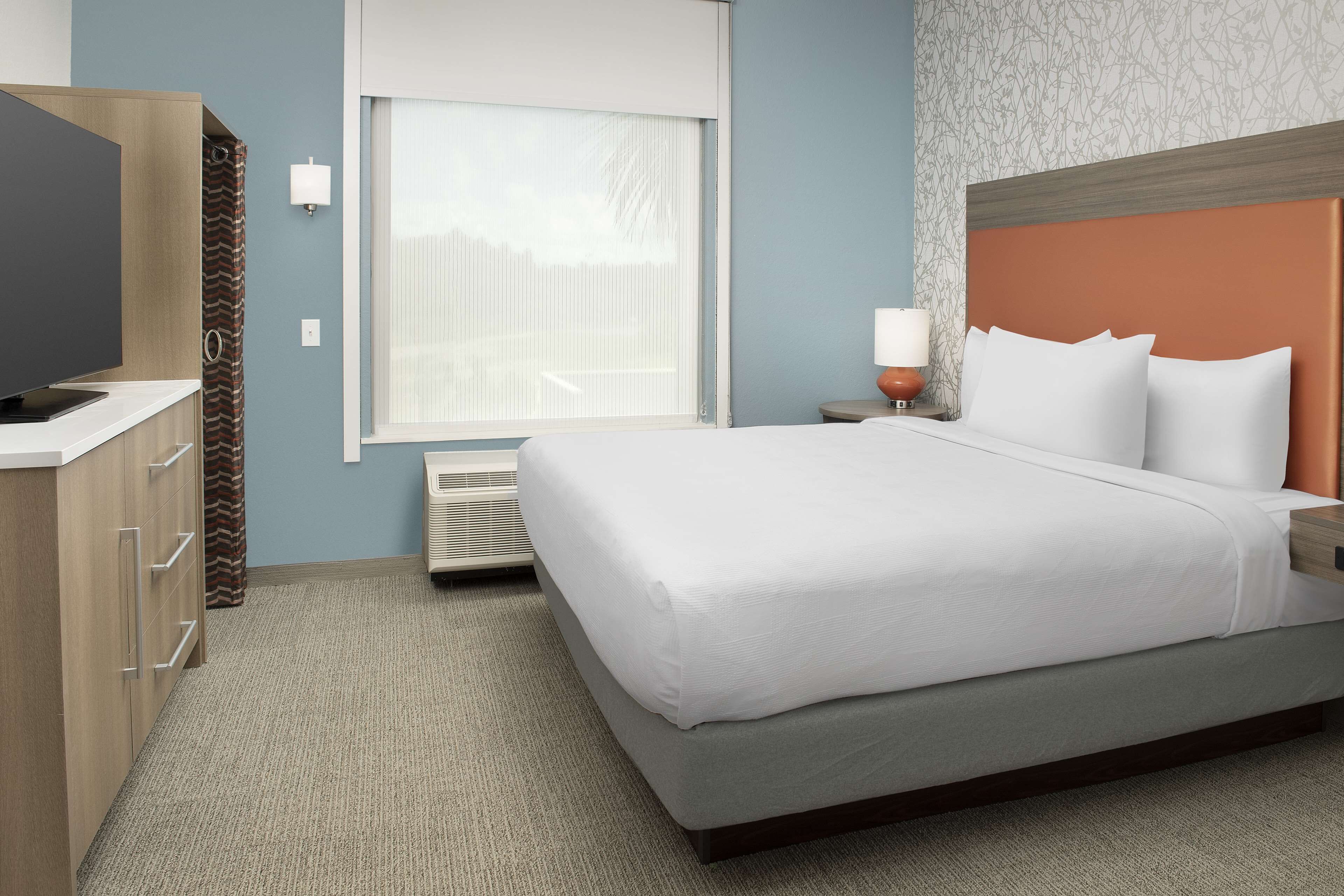A hotel room with a bed, a television, a nightstand, and a window with blinds.
