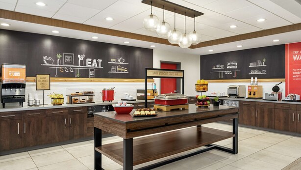 A modern breakfast area with a central table, kitchen appliances, and a wall with breakfast-related graphics. - Hampton Inn & Suites Olean (Olean)
