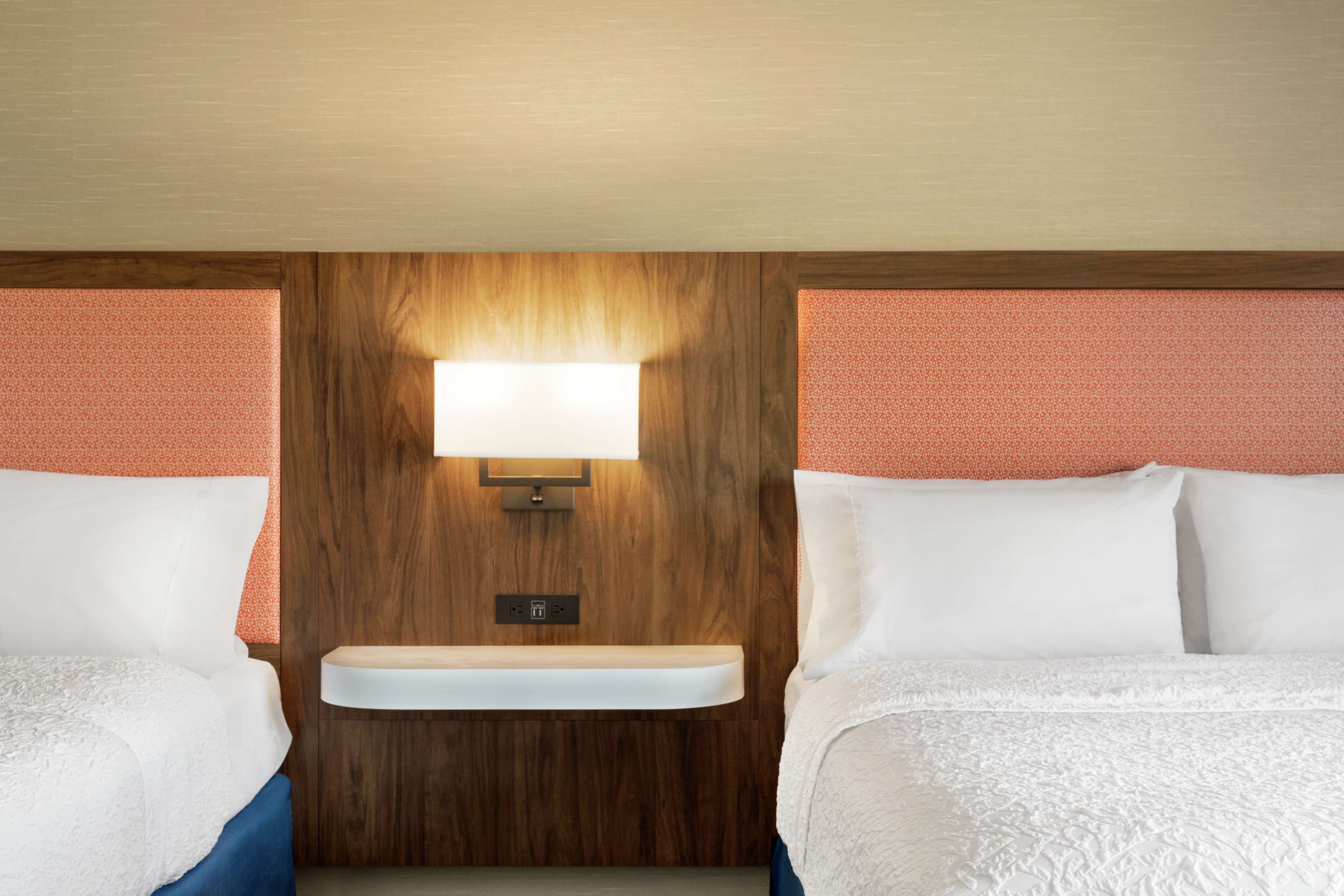 A hotel room with two beds, a wooden headboard, a built-in shelf, and a wall-mounted light fixture.