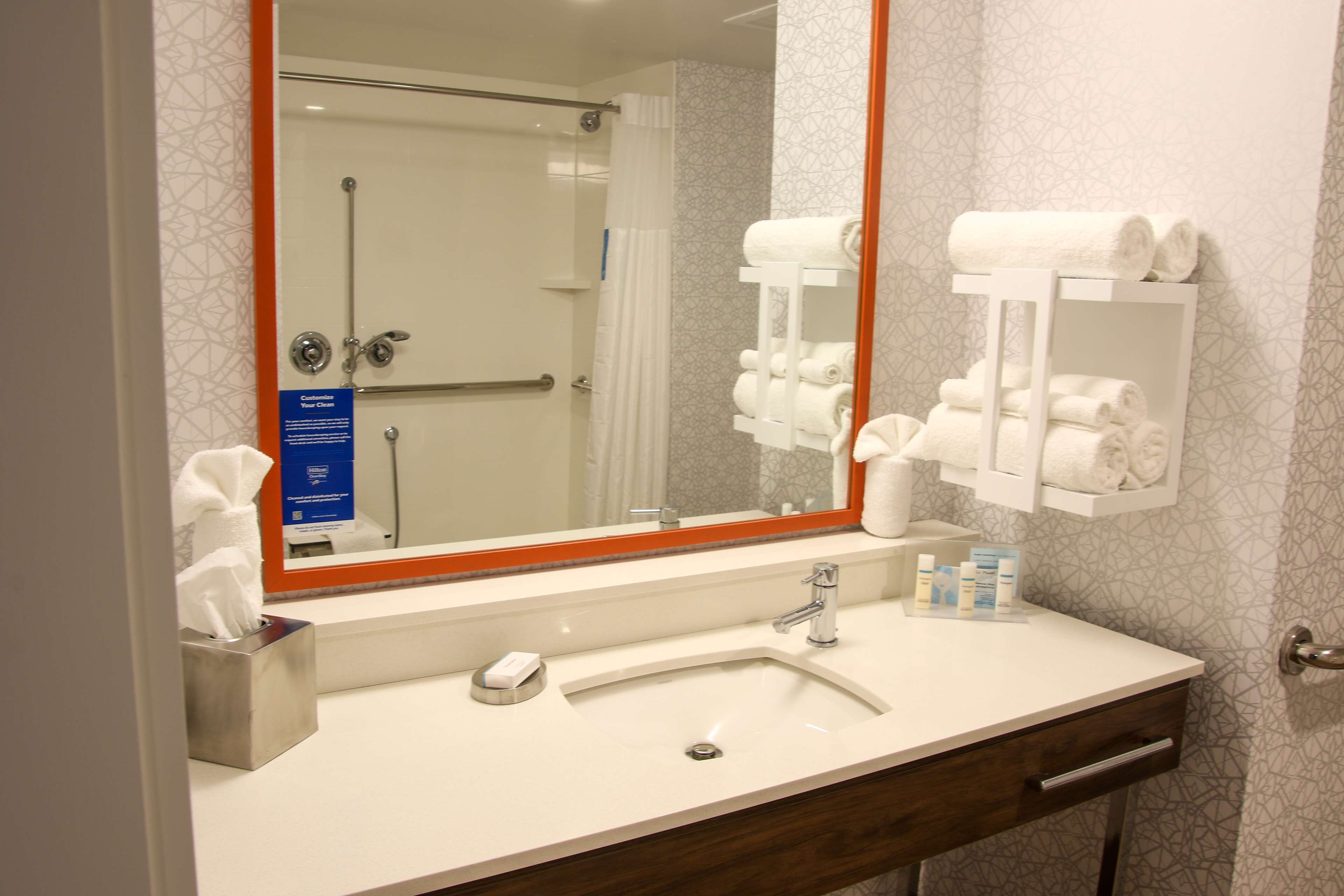 Un baño de hotel con lavamanos, espejo, toallero y puerta de ducha.