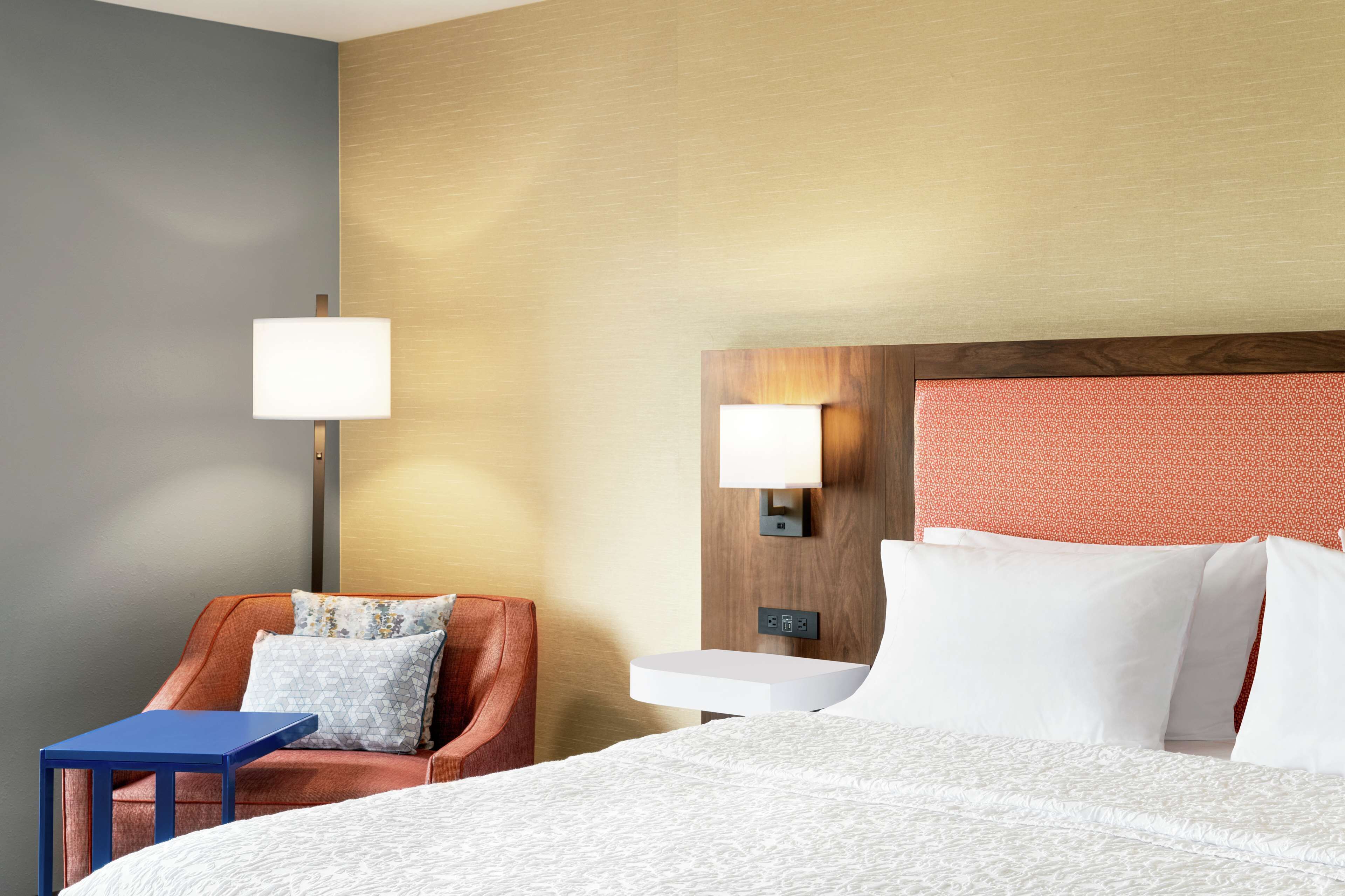 A hotel room with a bed, a nightstand, a lamp, a chair, and a small table.