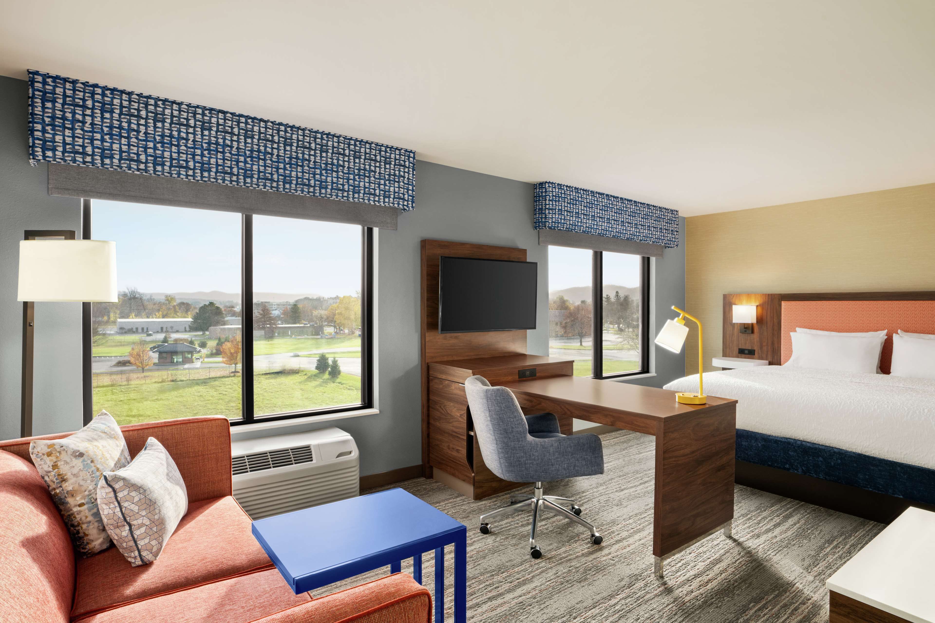 A hotel room with a desk, chair, bed, and a view of a park.
