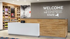 A hotel reception area with a wooden reception desk, a wall with a welcome sign, and shelves stocked with snacks and toiletries. - Hampton Inn & Suites Olean (Olean)