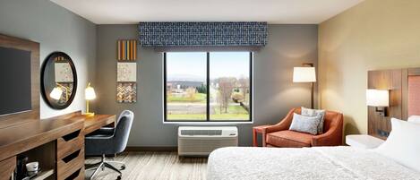 A hotel room with a bed, desk, chair, and a large window offering a view of the outdoors.