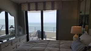 Un dormitorio con una cama grande, una mesita de noche, una lámpara, un espejo y vista a la playa.
