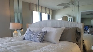 Una cama bien hecha con una colcha blanca y varias almohadas, una lámpara de noche y un televisor sobre una mesita junto a la cama.