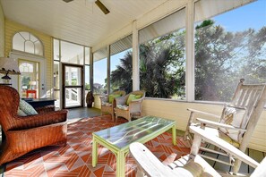 A sunlit room with wicker furniture, a glass-top table, and large windows offering a view of greenery.