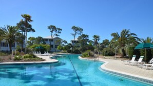 A clear swimming pool with lounge chairs and umbrellas, surrounded by palm trees and a building in the background.