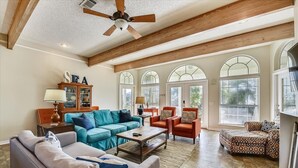 A spacious living room with a high ceiling, wooden beams, and large windows. There are multiple seating areas including a sofa, chairs, and a coffee table.
