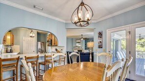 A dining area with a wooden table and chairs, a chandelier, and a kitchen in the background.