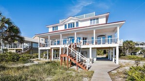 Une maison à deux étages située en bord de mer, dotée d’une terrasse en bois et d’un escalier menant à l’étage supérieur.