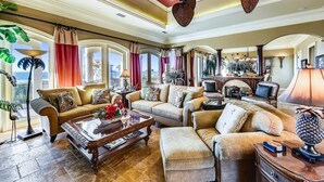 A spacious living room with a large sofa, a coffee table, and a fireplace. There are multiple windows with curtains, and a dining area is visible in the background. - Welcome to Deja Vu! (St. George Island)