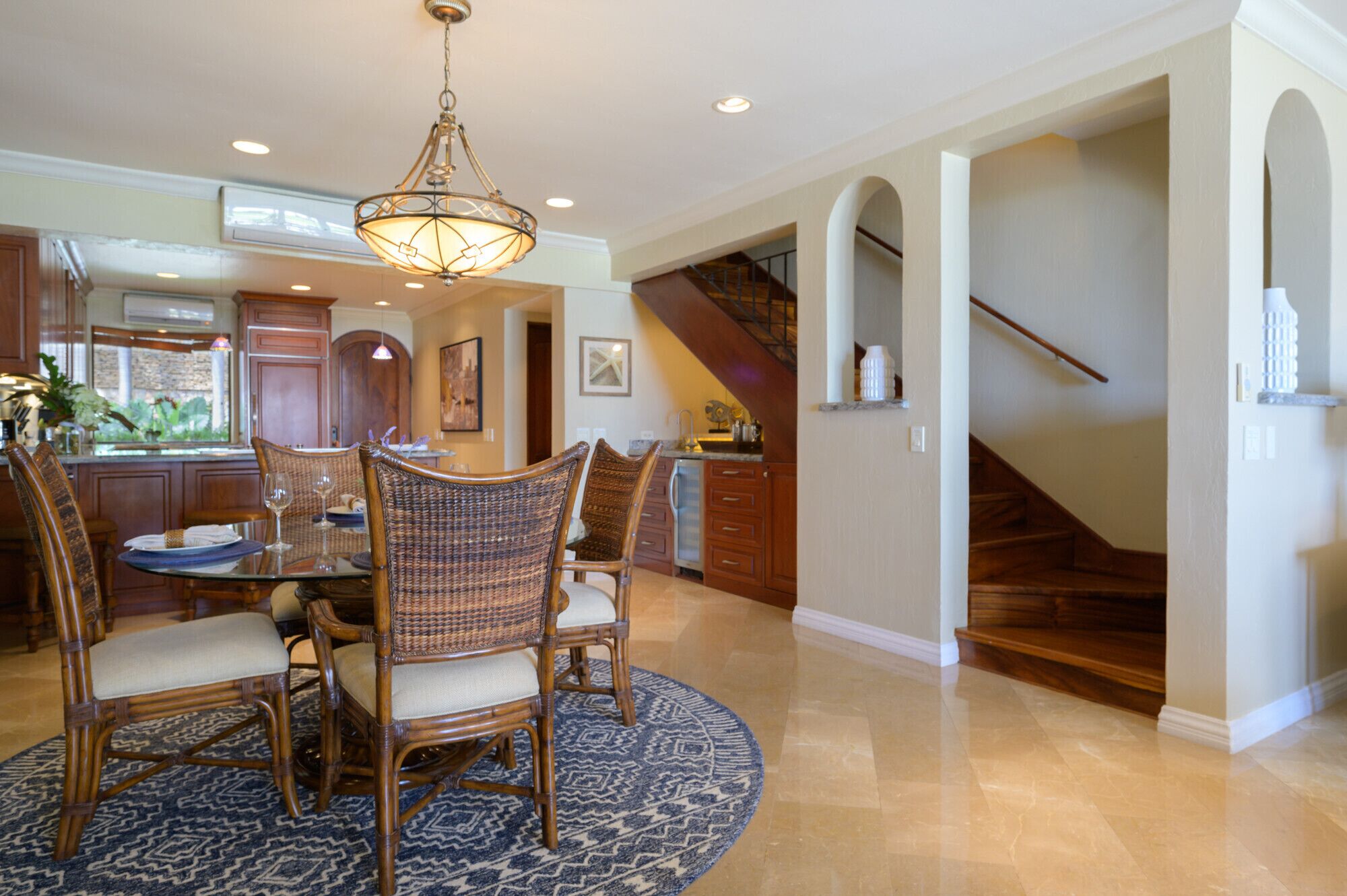 A spacious dining area with a round table and wicker chairs, a chandelier, and a view of the kitchen and staircase.