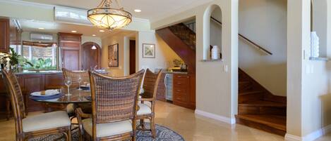 A spacious dining area with a round table and wicker chairs, a chandelier, and a view of the kitchen and staircase.