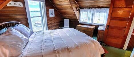A cozy, wooden cabin bedroom with a large bed, a small table, and a window overlooking a body of water.