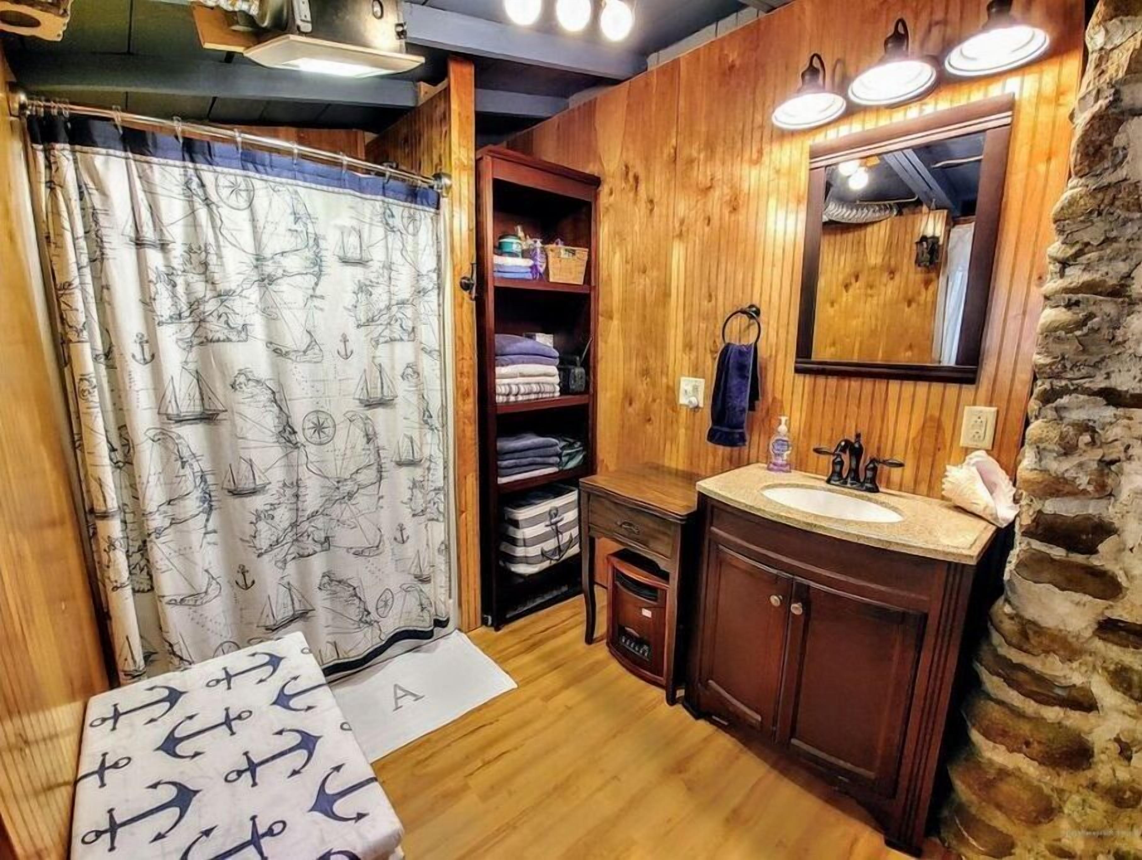 A wooden-themed bathroom with a nautical-themed shower curtain, a vanity with a mirror, and shelves with folded towels.