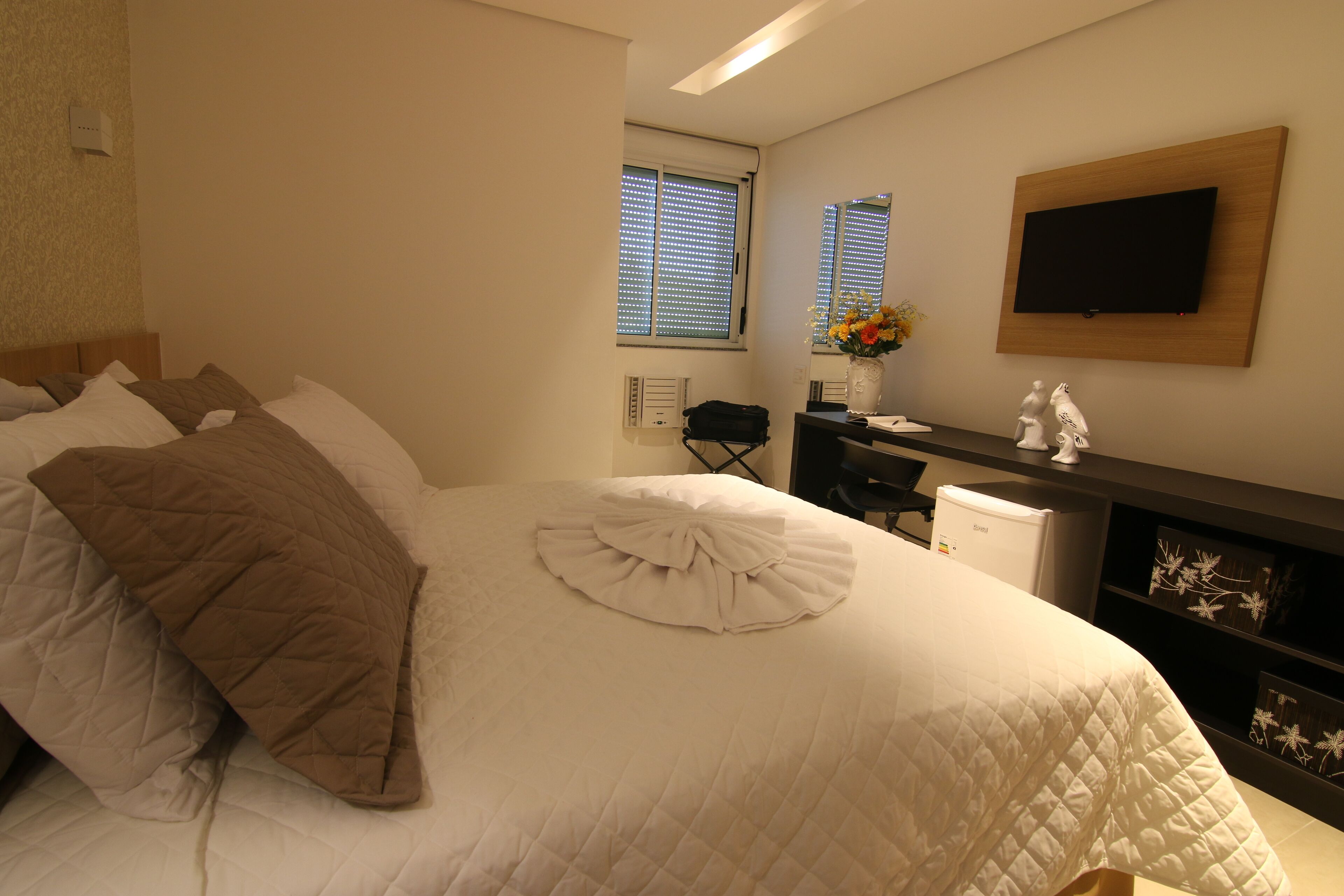 A neatly made bed with a quilted white duvet and a brown throw blanket, a flat-screen TV mounted on the wall, a small desk with a chair, and a vase with flowers on the desk.