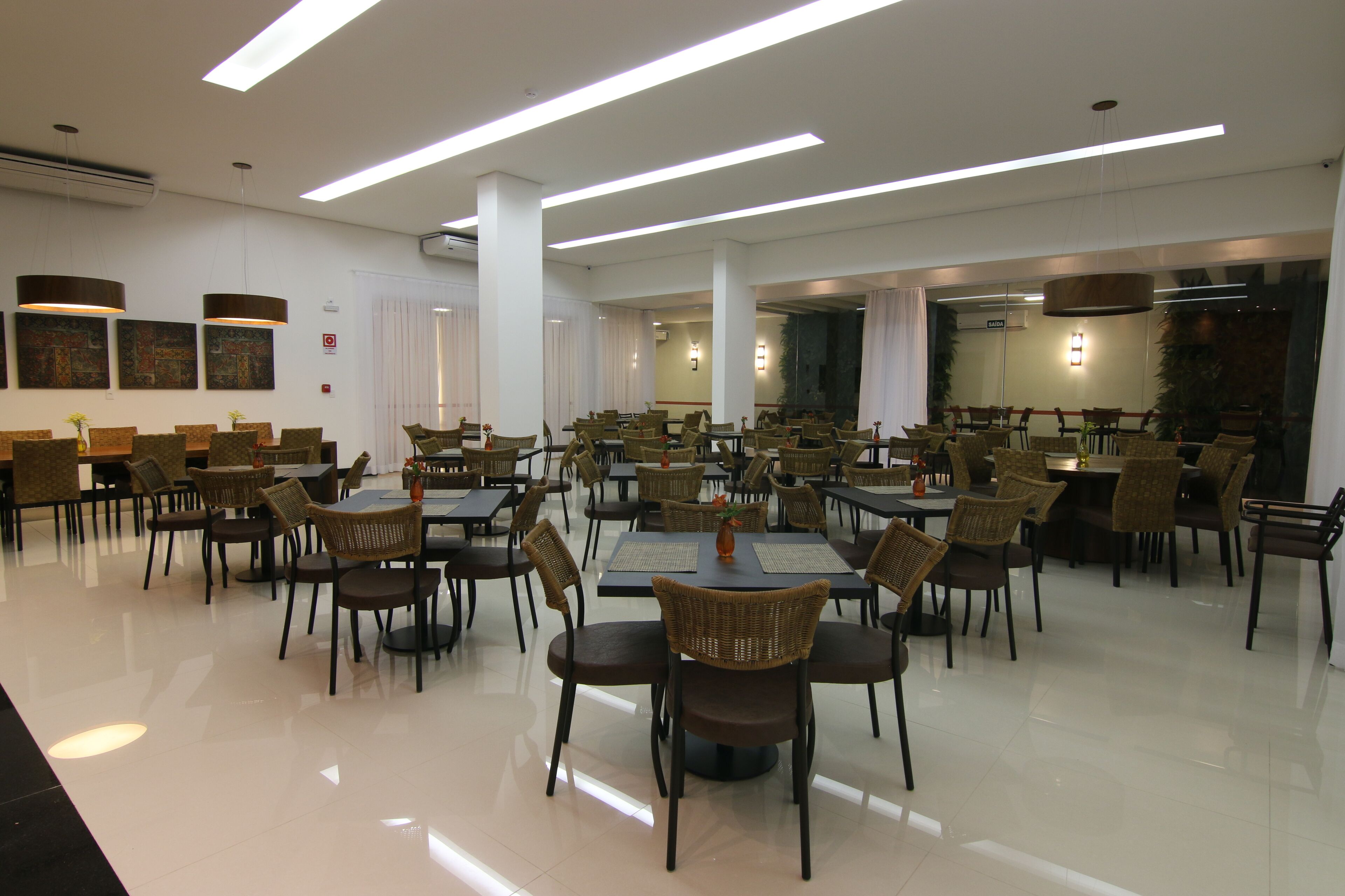 Uma sala de jantar espaçosa com várias mesas e cadeiras, grandes janelas e luminárias modernas.
