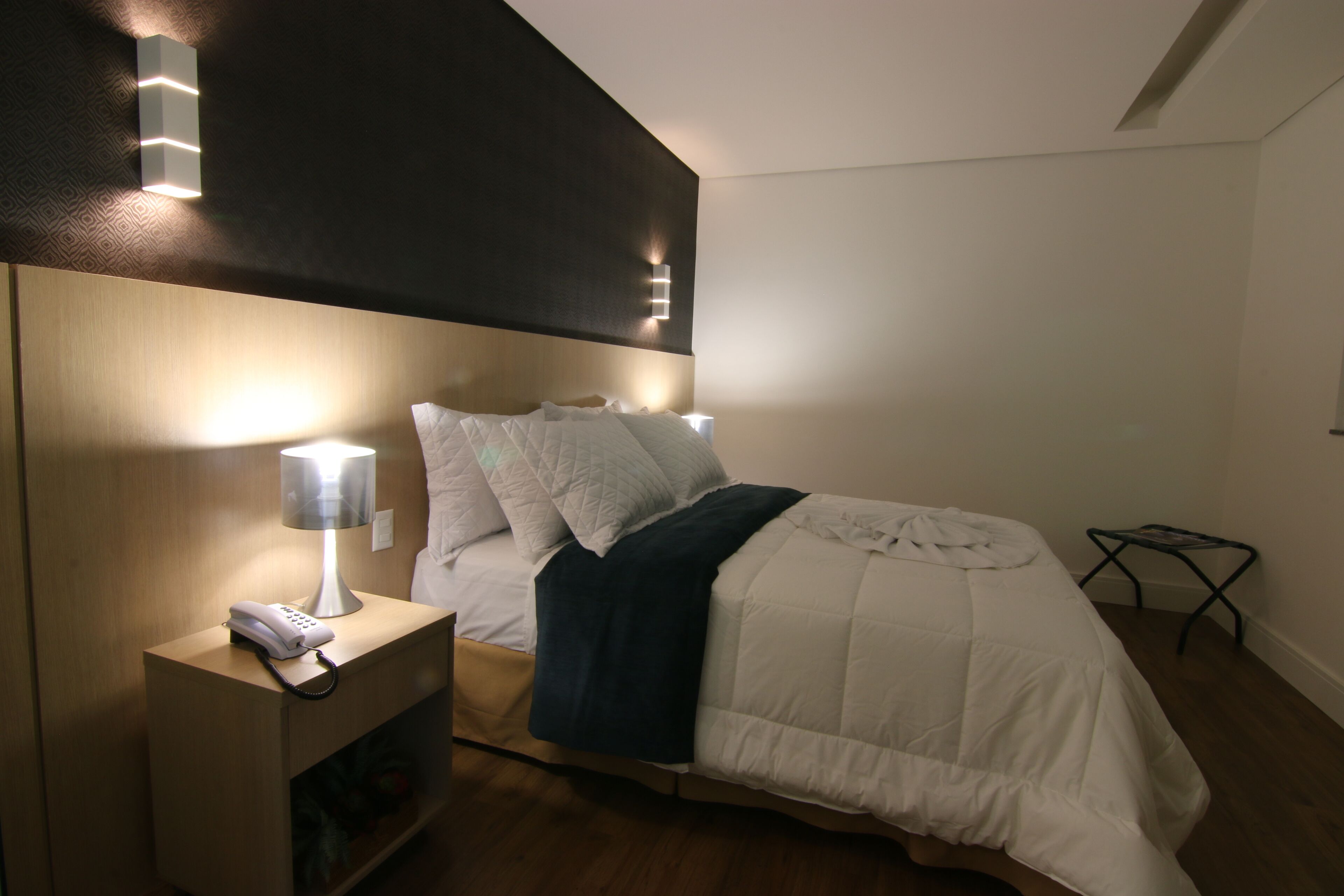A neatly made bed with a white comforter and pillows, a bedside lamp, and a telephone on a wooden nightstand.