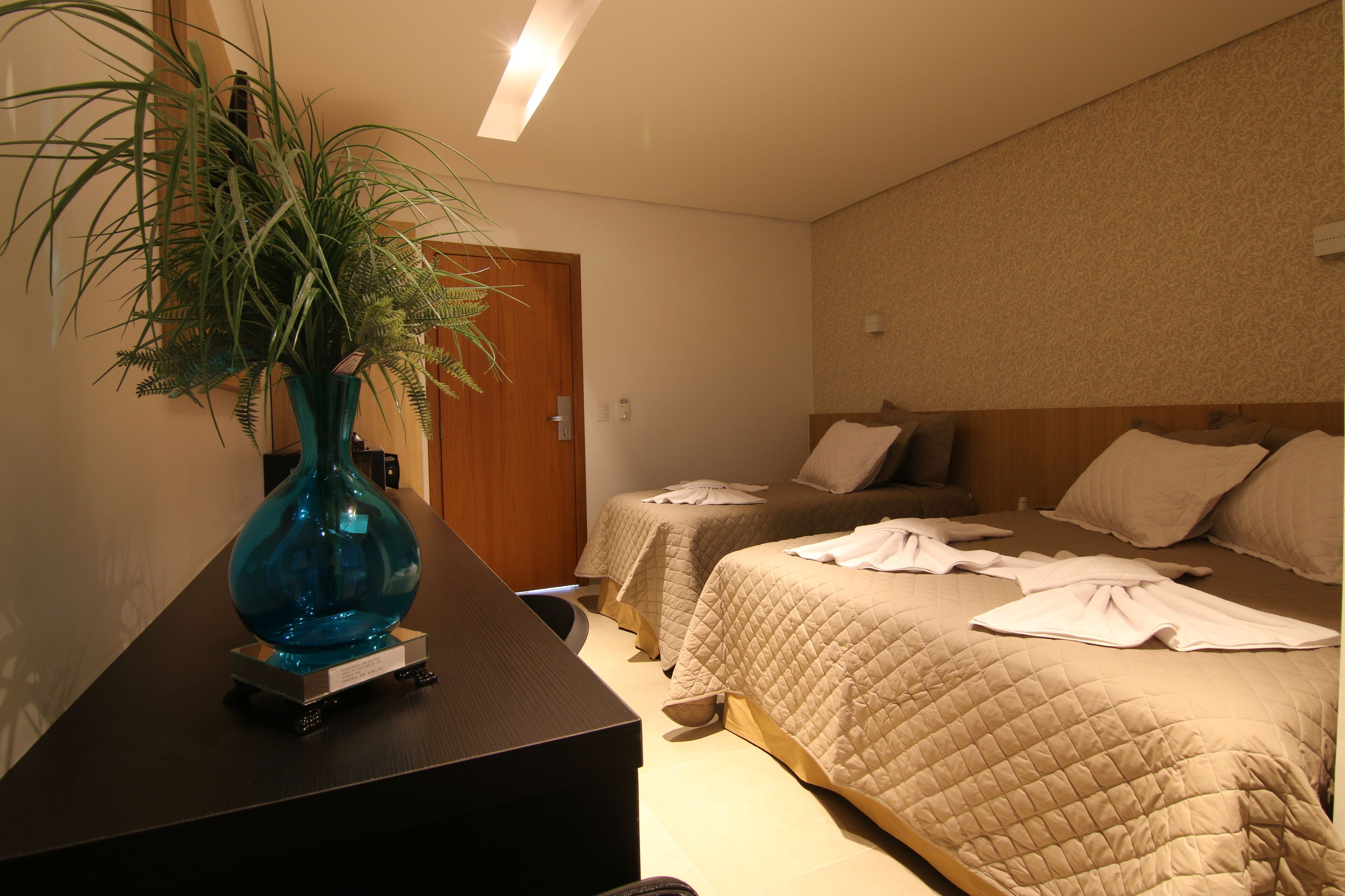 Quarto de hotel com duas camas, uma mesa de cabeceira com um vaso e uma porta ao fundo.