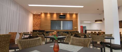 A modern dining area with multiple tables and chairs, a central vase with flowers, and a large screen on the wall.