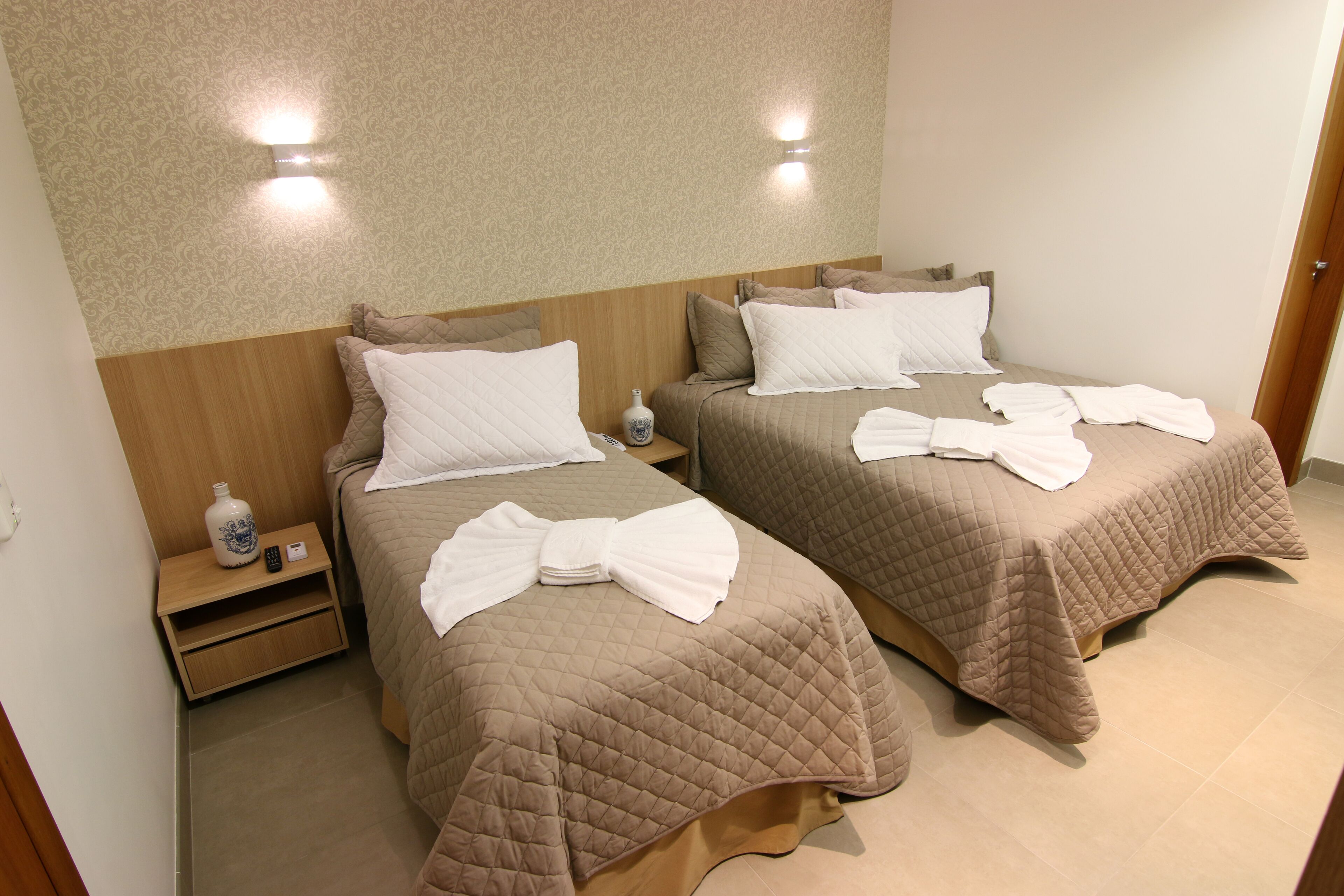 A hotel room with two beds, each with a brown quilted bedspread and white pillows. There is a small bedside table with a vase and a remote control.