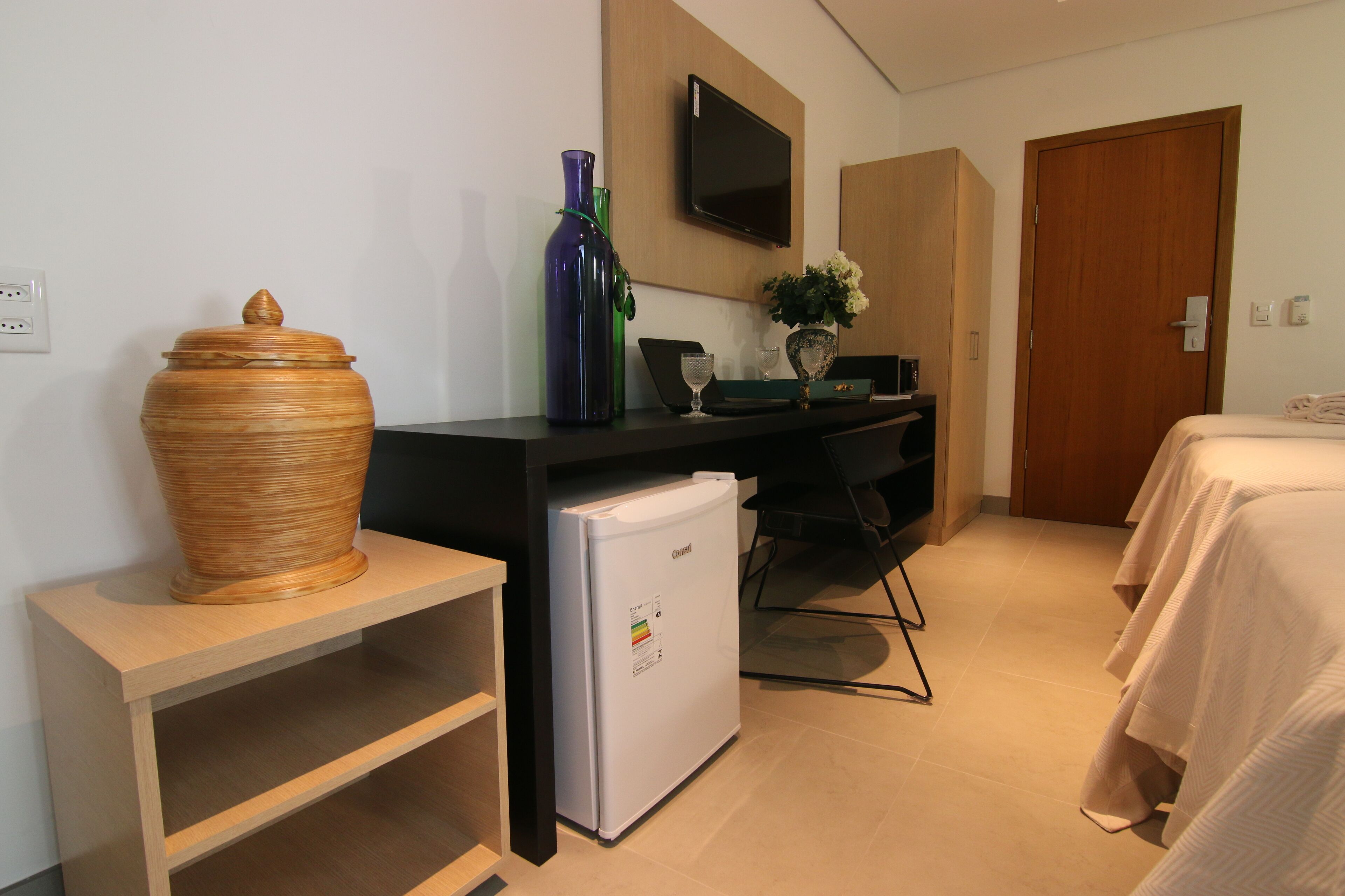 Ein Hotelzimmer mit einem Holznachttisch, einem weißen Kühlschrank, einem flachbildfernseher an der Wand, einer Holztür und einem Bett mit weißer Bettdecke.