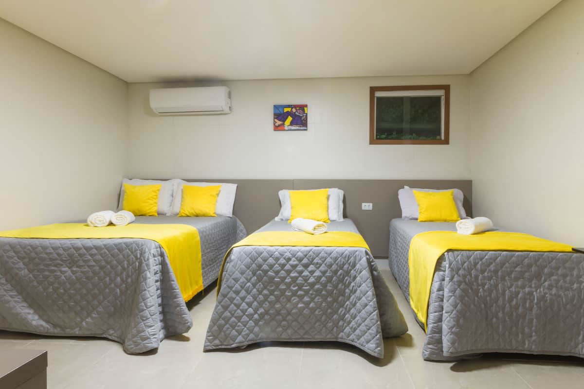 A hotel room with three beds, each with a quilted grey bedspread and yellow pillows.
