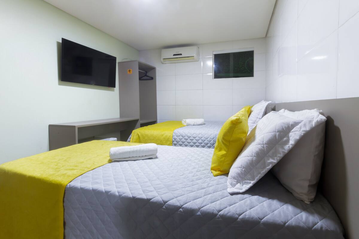 A hotel room with two beds, each with a quilted grey and white duvet, yellow pillows, and a white towel. There is a flat-screen TV mounted on the wall, a wardrobe, and an air conditioning unit.