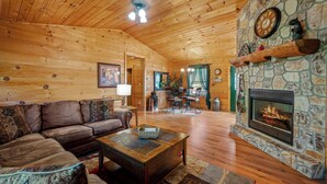 A cozy cabin interior with a stone fireplace, wooden ceiling, and a dining area in the background. - Quiet One-Level Cabin, Hot Tub, Fire Pit, WiFi, Parkway Close (Sevierville)