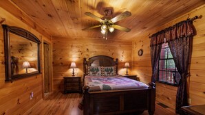A wooden cabin bedroom with a bed, two bedside tables, a mirror, a ceiling fan, and a window with curtains. - Quiet One-Level Cabin, Hot Tub, Fire Pit, WiFi, Parkway Close (Sevierville)