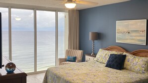 A bedroom with a large bed, a ceiling fan, a nightstand, a lamp, a chair, and a view of the ocean.