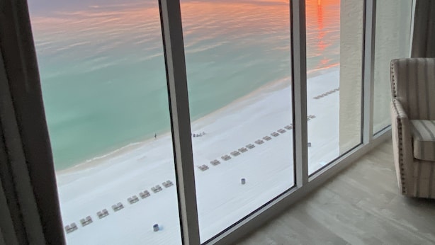 A room with a large window overlooking a beach at sunset.
