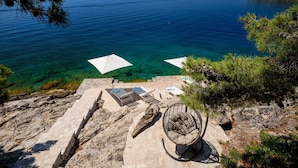 Une terrasse en pierre avec des espaces de détente en plein air, comprenant des chaises longues et des parasols, offrant une vue sur une mer d’un bleu limpide.