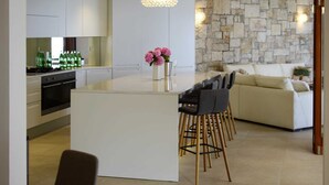 Une cuisine moderne avec des meubles blancs, un îlot central et des tabourets de bar. On aperçoit un mur en pierre au fond et un canapé beige dans l’espace salon.